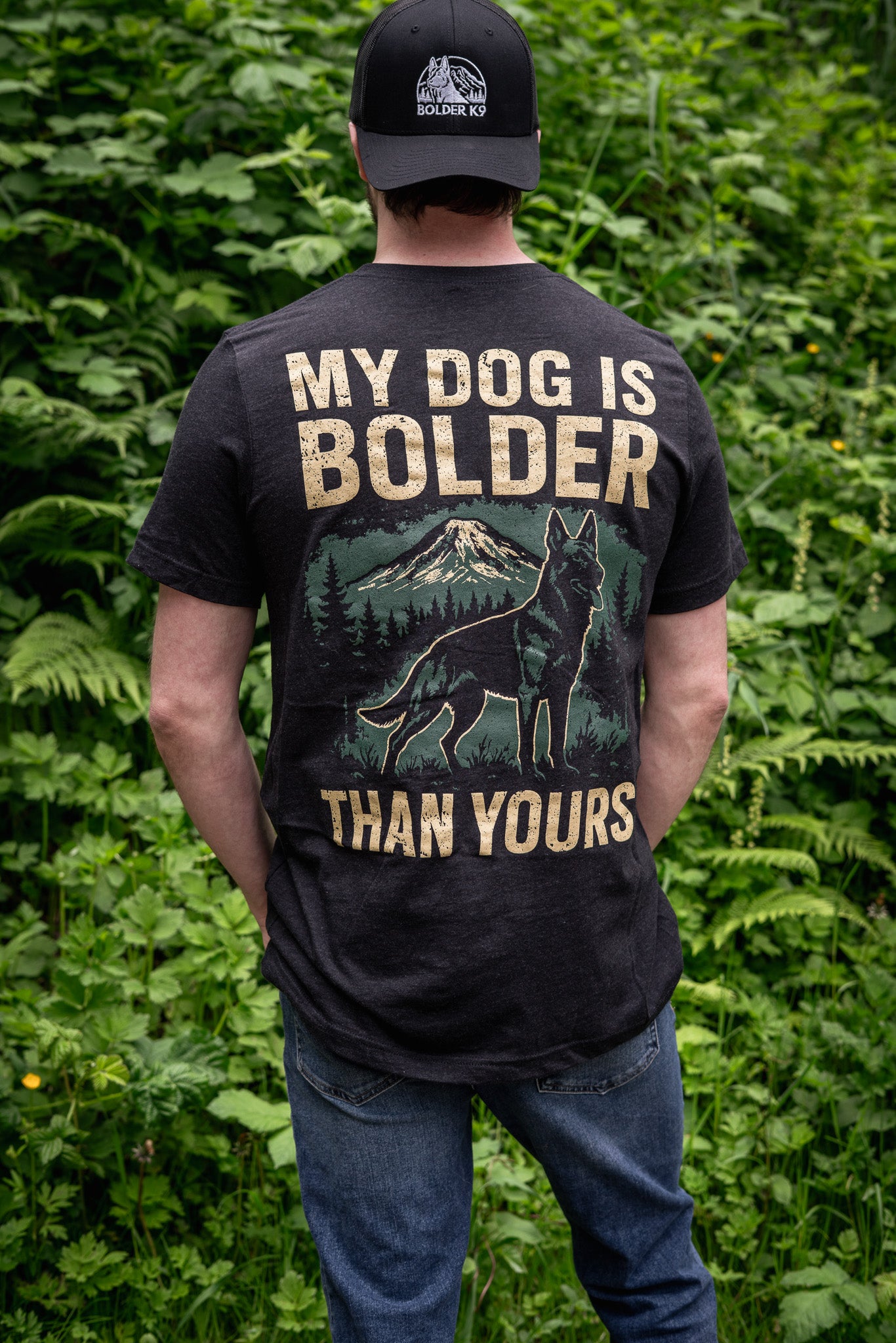 Man wearing 'My Dog Is Bolder Than Yours' t-shirt in heather black against a green foliage background.