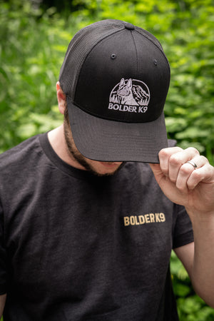 Man wearing Bolder K9 trucker hat and shirt, surrounded by greenery, showcasing outdoor style and comfort.