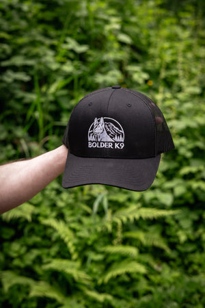 Bolder K9 trucker hat held in hand, featuring bold logo, surrounded by lush greenery.